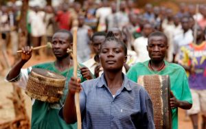 Civilians engage in a march in the Kibondo region. They are marching while playing the drums and are walking down are narrow street. There are onlookers on either side of the street.