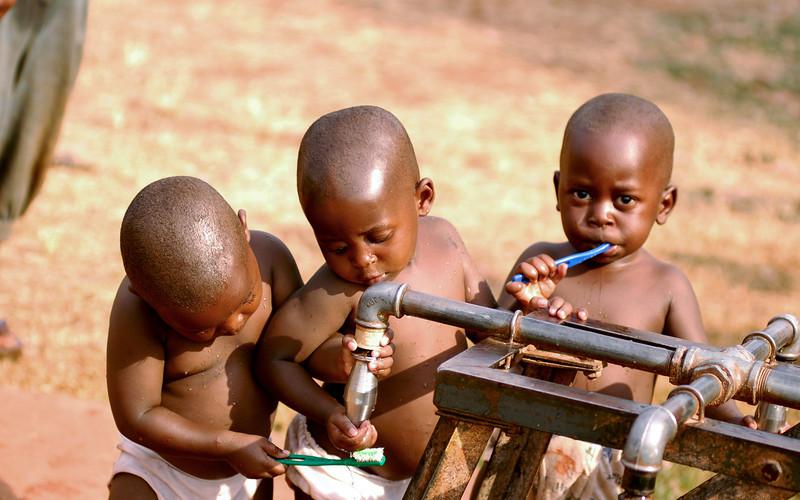 Three young children are shown at a water faucet and are brushing their teeth.