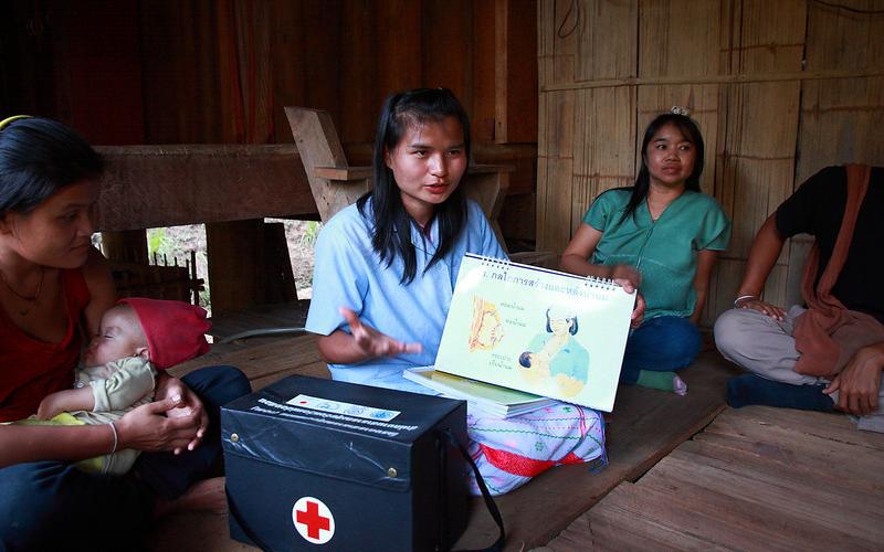 A young woman sits in a wooden house surrounded by three other women. One of the women is carrying a child. There is a medical kit next to the woman speaking, and she is holding a diagram of a mother and her child.