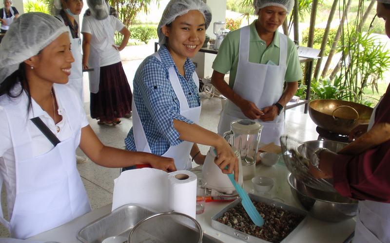 Three young girls stand at a white table and prepare food. There are cooking utensils and bowls on the table. The girls are wearing hair nets and aprons and there are several girls standing in the background.