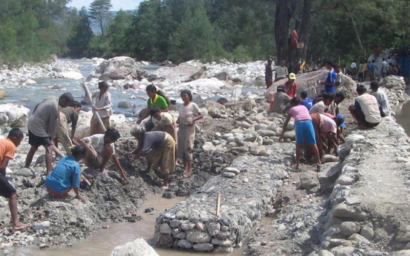 A group of people sit on beds of rock and concrete next to a running river. They are all digging into the stone. There are trees in the background.