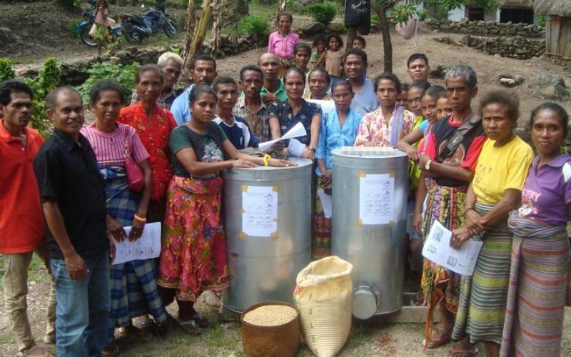 A large group of individuals stand on a hill surrounded by dirt, trees and huts. They are standing with two metal containers and bags of rice.