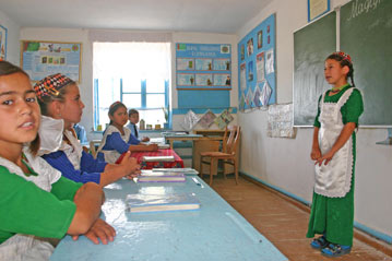 Three girls wearing traditional clothing sit on wooden chairs in front of a computer and a projector. There are older men and women standing around watching them. There are posters hanging on the wall.
