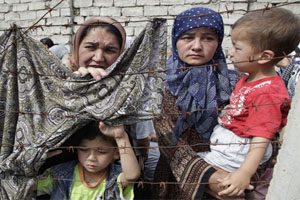 Two women holding young children peer through a barbed wire. There is a blanket caught on the barbed wire.