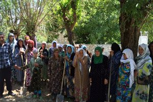 A dozen women stand with shovels in a field. There are large trees lining a stone wall.