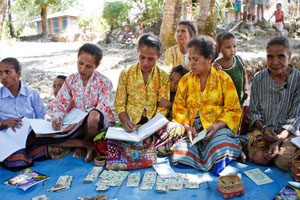 A group of young women sit on a blue plastic sheet. They are using pens to write in books. There is money on the floor in front of them.