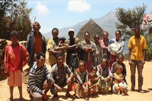 A group of men, women and children stand in a dirt paddock with their arms folded. One woman is holding a baby in a sling. There is a hut and mountain in the background.
