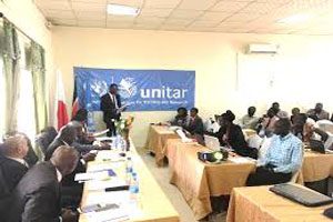 A man stands at the front of a room reading from a sheet of paper. Several people are seated at desks in front of him with orange table covers. Behind the man addressing the room is a sign stated ‘unitar.’