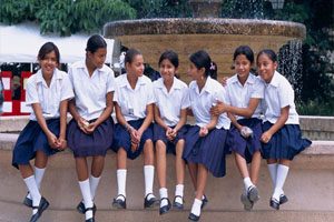 Seven girls sit on the edge of a large fountain. They are wearing white shirts and blue skirts. There are trees in the background