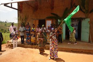 A woman is standing outside a brown house and is waving a green flag. Behind her, a small crowd has gathered out the front of a hut and the onlookers are cheering her on.