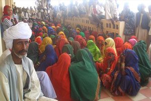 A large crowd of women in colorful dresses are gathered outside a building on the floor. Their bodies are covered in traditional dress except for their eyes. More people are seated on top of a brick wall.