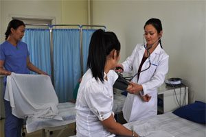 A female doctor wearing a white lab coat with a stethoscope checks the blood pressure of a young Mongolian woman sitting on a bed. There is a nurse behind them wearing a white uniform and she is folding a sheet.
