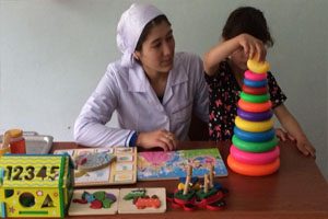 Woman sits at a wooden desk with a child. There are games for children on the desk, and a child is playing with a tower shaped toy.