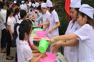 Girls dressed in white clothing with white hats stand at desks and wash the hands of smaller girls using bright green and pink plastic bowls and cups.