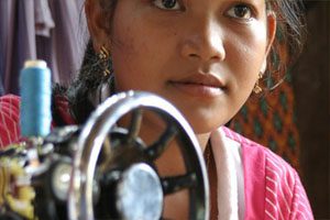 A young woman stares into the distance and is wearing a stripy pink and white top. She is sitting behind a sewing wheel.