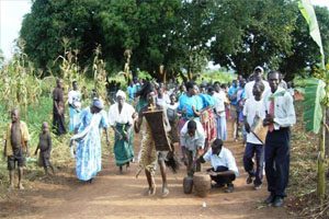 A large group of people are dancing in a field. Two men are playing the drums, while others are clapping enthusiastically.