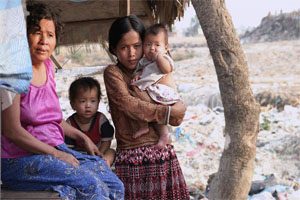 Two women stand under a tree and a hut in the middle of a field. They are holding two young children.