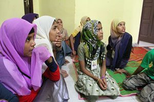 Eight young women sit on a tiled floor with a rug. They are looking to the right.