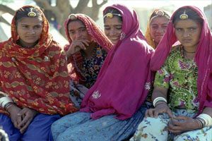 Five women sit together and pose for the camera. They are wearing headpieces and colourful scarves.