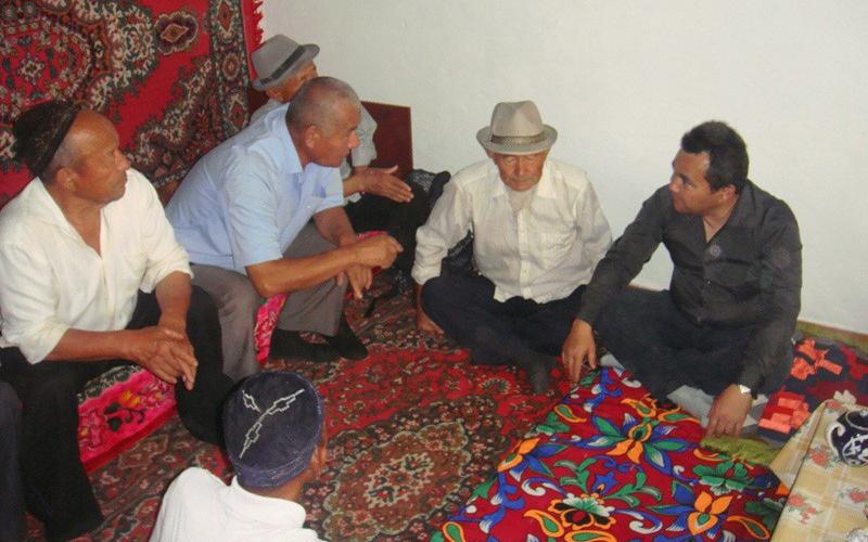 A group of six men sit on colourful rugs in a white room, with their legs crossed.