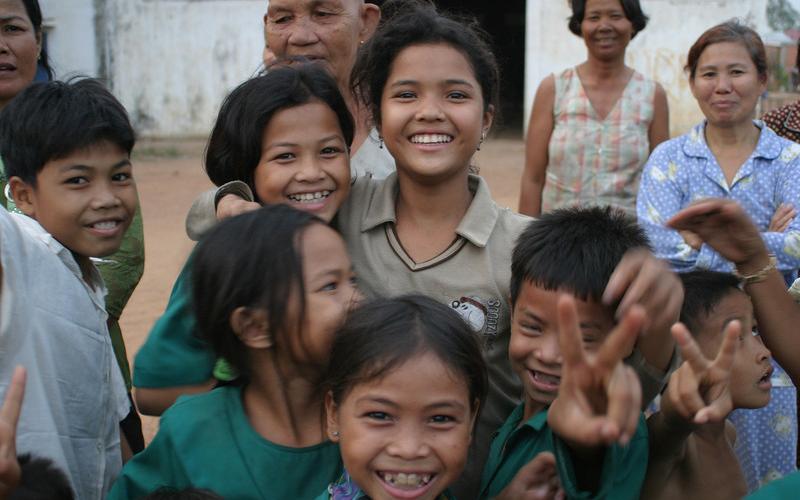 A group of young boys and girls smile in front of a camera in a dirt field. Two of the children are doing a ‘peace’ sign with their hands. There are older women standing in the background watching the children.