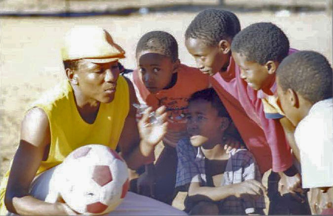 A young man holding a soccer ball in one of his hands gathers a small group of boys, who are attentively listening to him.