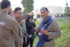 Local authorities stand in a field in Lebanon. One man, on the right, is wearing a vest and a radio transceiver. A woman behind him is taking a photo of the field with a large camera.