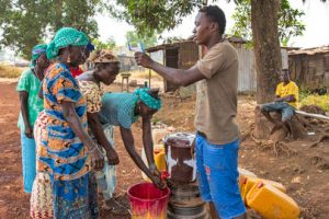 A group of people are standing next to canisters full of water. A woman is washing her hands underneath a stream of water.