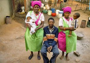 A man sits on a chair surrounded by two older women who are carrying children.