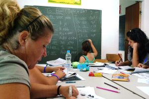 Four women sit at a large desk scattered with crayons, pencils and paper. A woman writes on the chalkboard in front of the table.