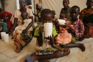 A group of young children sit on the floor on a piece of fabric. They are sipping from large white cups. There are pieces of fabric hanging on the wall behind them.