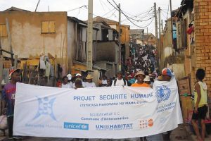 A large group of people walk down a narrow street carrying a sign stating ‘Projet Securite Humaine.’ There are houses lining the street.