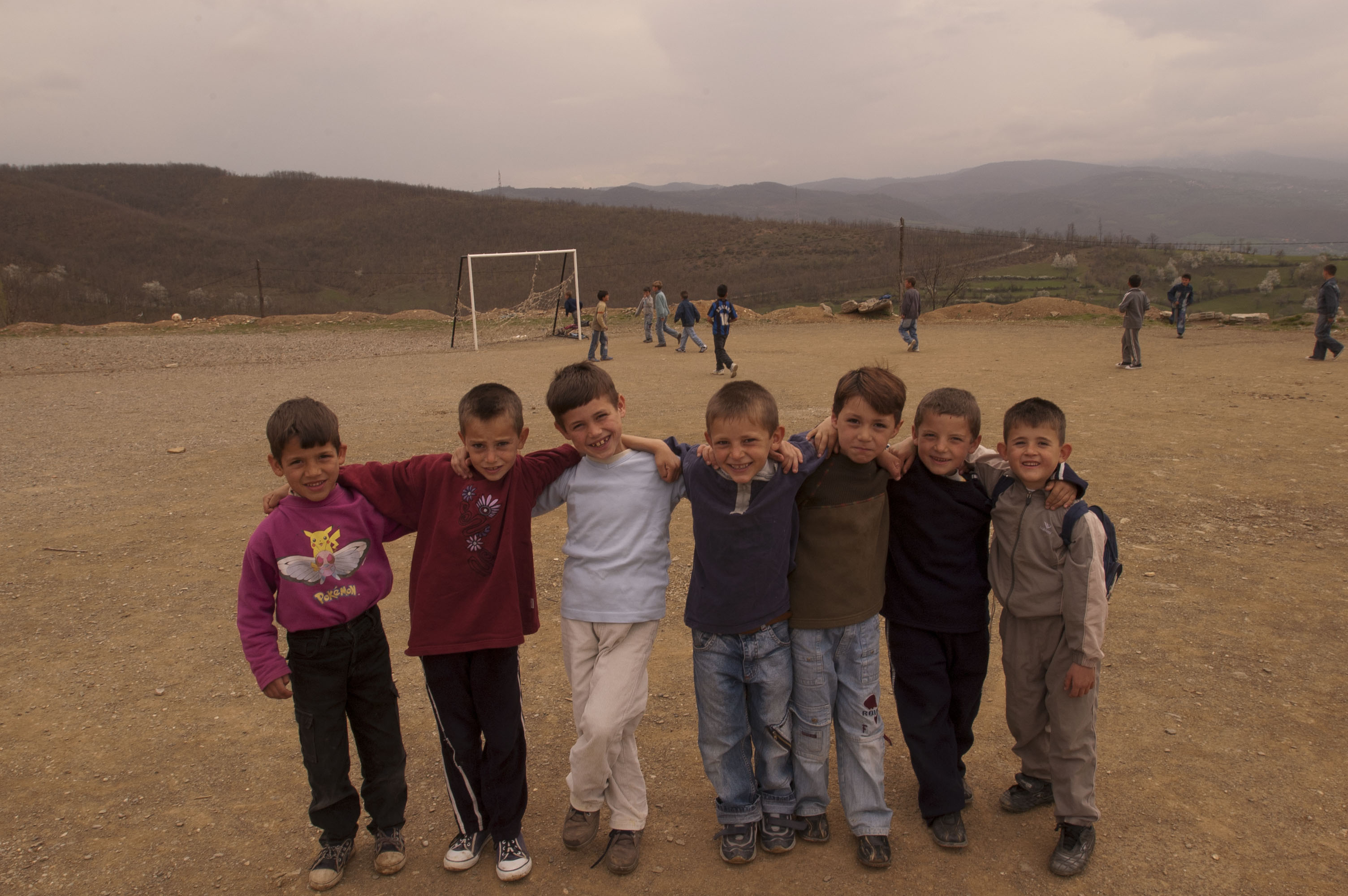 Seven young students stand at the camera smiling, with their arms around each other’s shoulders. They are standing in a dirt soccer field, and there are young boys playing behind them near a soccer goal. There are mountains in the backdrop.