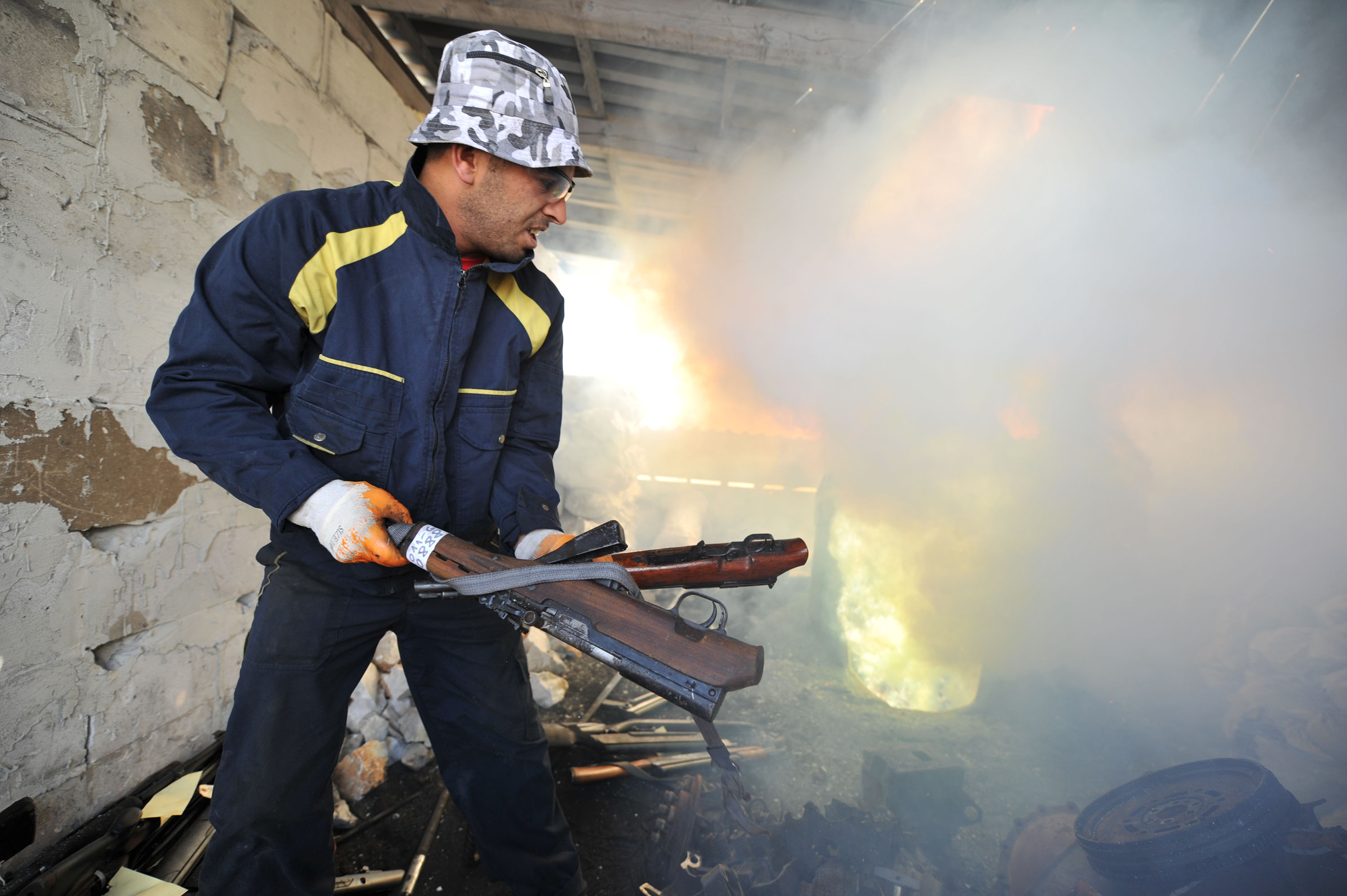 A man wearing long sleeved pants, shirt, gloves, a hat and sunglasses holds two guns and is about to throw them into a fire as part of a gun destruction programme.