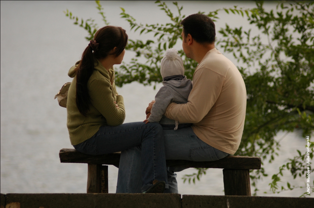 A woman and man sit on a park bench with their legs crossed together. The man has a child on his lap who is wearing a beanie. They are looking out into the distance and there is a tree in the background.