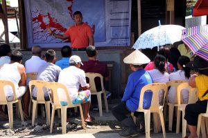 A man stands in front of a map and instructs a group of men and women sitting in plastic chairs. There are women carrying umbrellas for shade.