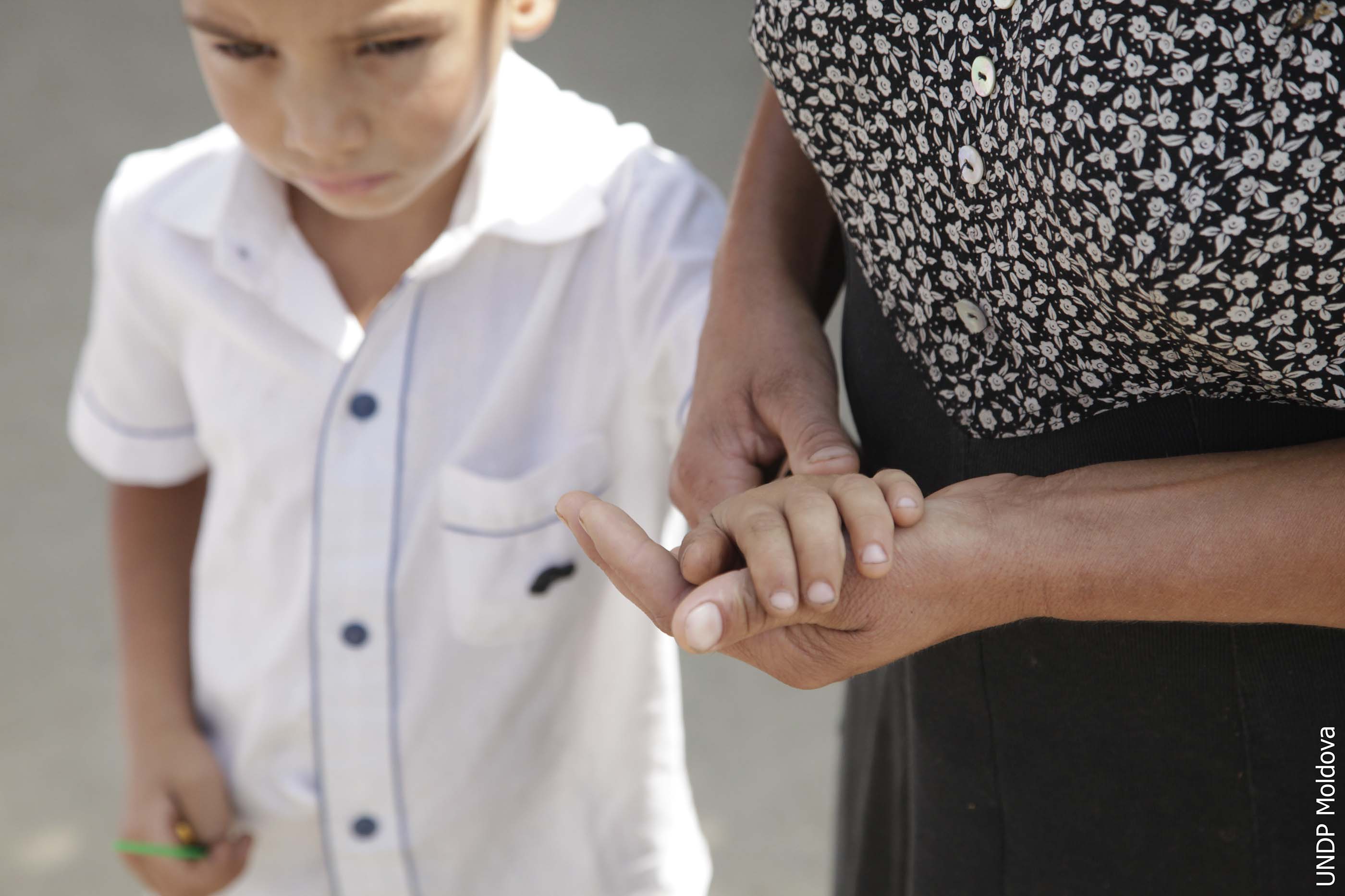 A child looks to the floor as a woman next to him holds his hand. The head of the woman is not visible in the photo.