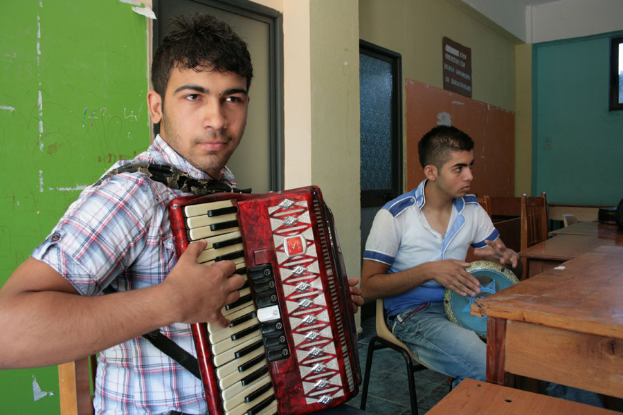 Two young men play a drum and an accordion while sitting at a wooden table.