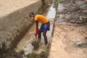 A man stands in a well which is full of mud and water. He is wearing a mask and is holding a shovel. He is digging mud out of the well.