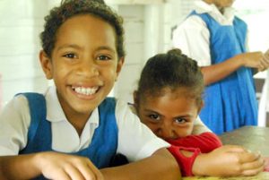 Two children smile in front of the camera as they sit at a wooden desk. Behind them is a brick wall that has been painted white.