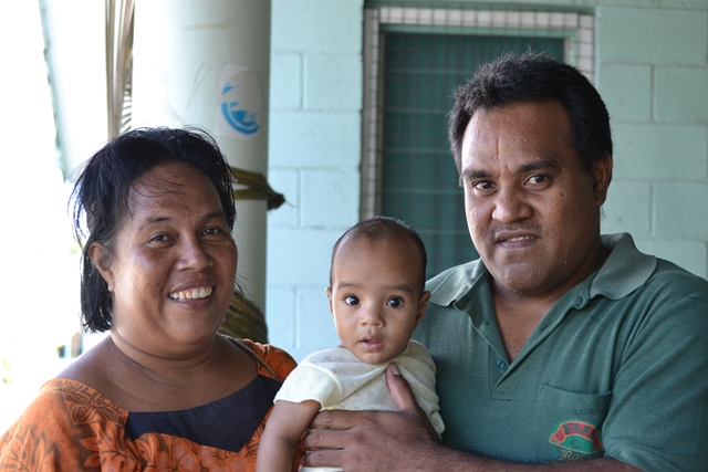 An adult woman and male smile in front of a camera and are holding their child. Behind them is a brick wall painted blue.