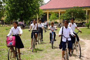 A group of children ride their bikes towards the camera. They are in a large green field. Behind them are trees and a yellow building with a tiled roof.