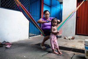 A woman sits in a hammock and brushes the hair of a young child standing in front of her. Underneath them is a concrete floor. There is a baby doll, bucket and pairs of shoes on the floor.