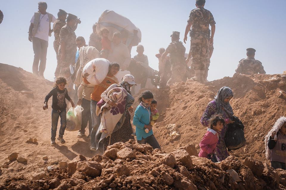 A group of adults and children walk down a dusty hill covered in rocks. They are carrying personal belongings and large white bags. There are members of the army watching the people as they move down the hill.