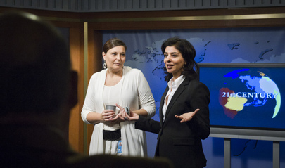 Two women stand in a television studio. One woman is wearing a black blazer and has her hands in an open gesture. There is a television behind them which is showing the text ‘21st Century.’