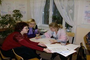 Three women sit around a wooden table. There are documents on the desk and a poster hanging on the wall. One woman is drawing on a sheet of paper spread out on the desk.