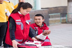 A young man sits down next to a young girl, who is holding a first aid kit. They are both wearing red vests. There are three people standing behind them in yellow shirts.