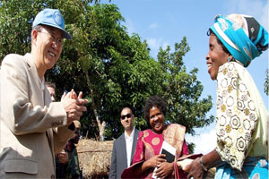 Ban Ki Moon stands next to a group of people watching a woman on the right. She is smiling and is holding a cob of corn. There is a bale of hay behind them and a large tree.