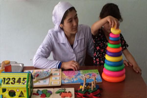 A woman sits at a table next to a young girl, who is playing with a cone shaped toy. There are other children’s games on the table.