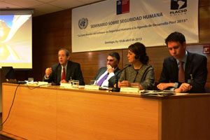 A panel of speakers sit at a wooden desk on a stage. There is a screen and a projector next to them. Behind them is a sign which states ‘Seminario Sobre Seguridad Humana